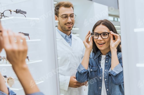 Preview: attractive smiling woman choosing eyeglasses and looking at mirror while male optometrist standing