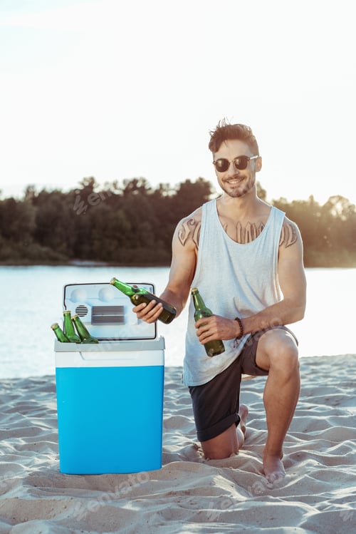 Preview: young smiling man taking beer from portable fridge on sandy beach