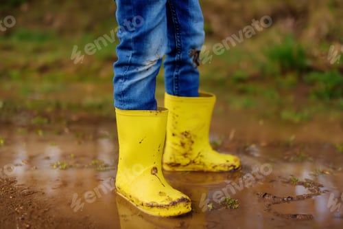 Preview: Mischievous preschooler child wearing yellow rain boots jumping in large wet mud puddle.