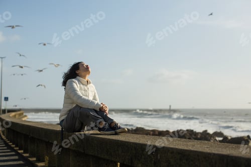 Preview: Beautiful Mixed-Race Asian Woman Sitting Of Sea Promenade. Seagulls Fly Over The Foaming Surf.