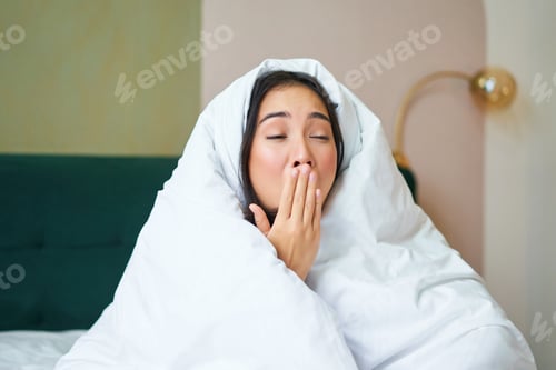 Preview: Close up of funny sleepy girl, asian woman yawns after waking up early morning, covers herself with