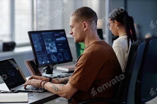 Preview: Profile of Focused Male Programmer with Septum Coding on Laptop at Working Desk in Office