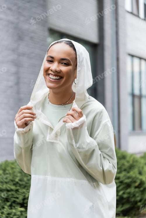 Preview: smiling african american woman in waterproof raincoat standing under rain outdoors