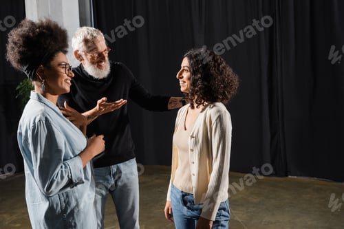 Preview: smiling art director pointing with hand and talking to multiracial woman near african american