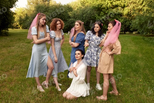 Preview: Happy charming bride in white dress with white veil sitting on the grass and bridesmaids with pink