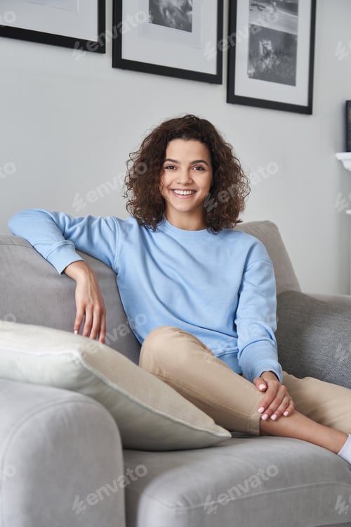 Preview: Smiling young pretty hispanic woman looking at camera sitting on sofa at home.