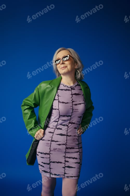 Preview: positive senior lady in trendy sunglasses and green leather jacket over purple dress standing with