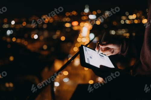 Preview: Close-up of man standing on balcony with urban view and using tablet at night