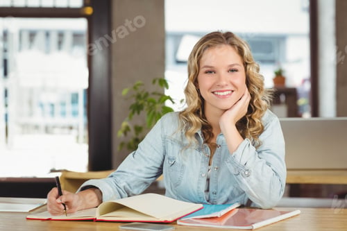 Preview: Female teenager smiling while writing in open notebook at wooden desk with laptop and pen