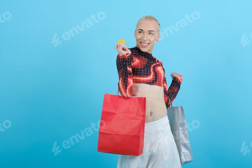 Preview: Young non-binary person holding a credit card and shopping bags while posing