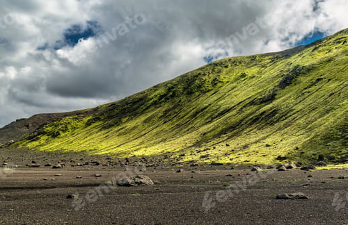 Preview: Vibrant green moss-covered hill under a dramatic cloudy sky in Iceland.
