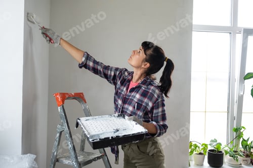 Preview: Woman in paint roller and white paint paints the wall of house on stepladder