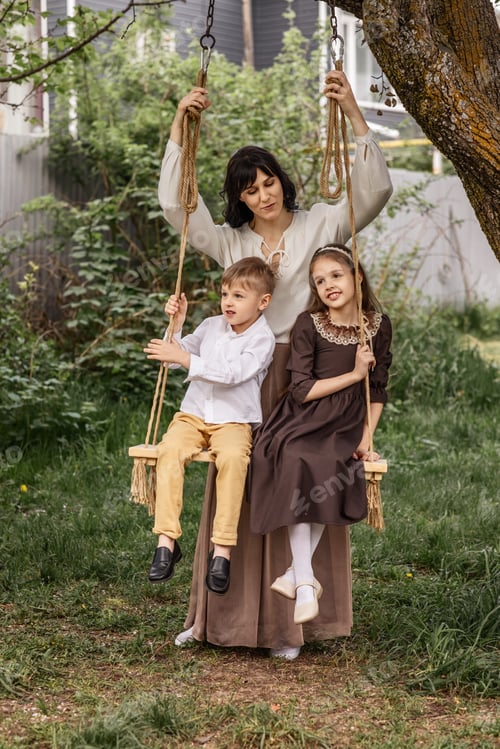 Preview: mom hugs two children sitting on a rope swing in the courtyard of a country house