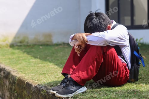 Preview: Sad young schoolboy sitting with hands on knees and head between his legs.