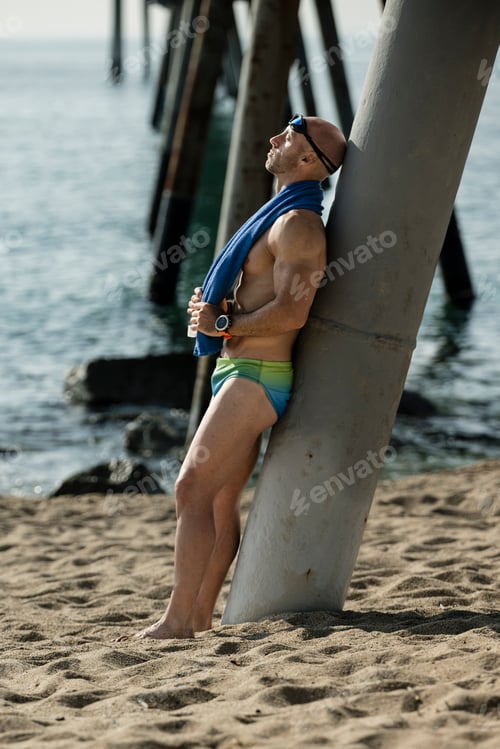 Preview: Professional swimmer toweling off his head with swim glasses sitting at the beach. Fit man with a