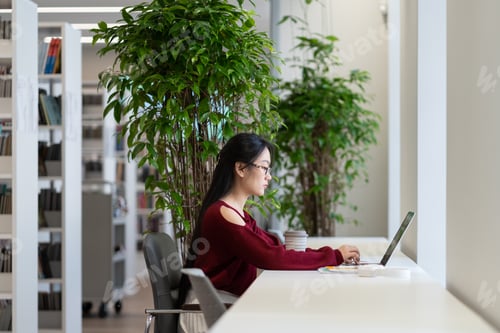 Preview: Focused Asian girl student using laptop while studying in university library, plant on background