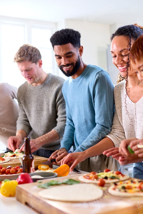 Preview: Group Of Friends At Home In Kitchen Adding Toppings To Homemade Pizzas For Party Together