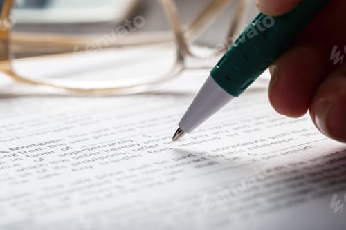 Preview: Close-up of a person writing with a green pen on a document with glasses nearby
