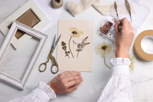 Preview: Woman putting dry flowers onto paper sheet at white marble table, top view