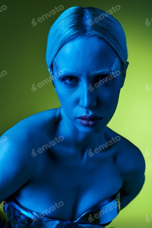 Preview: extravagant woman in robotic attire posing in blue lights on green backdrop and looking at camera