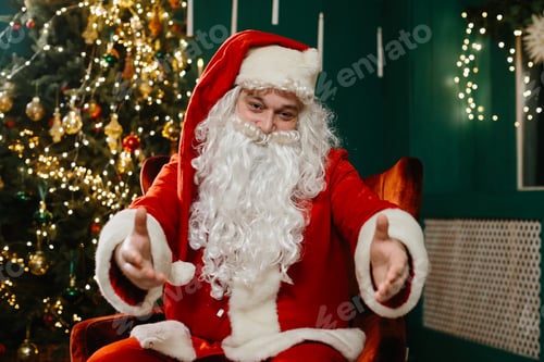 Preview: Santa Claus with outstretched arms looking at the camera, by the Christmas tree.