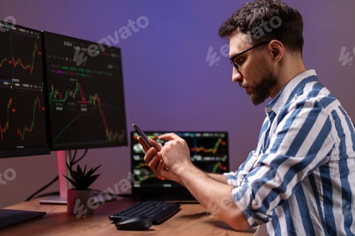 Preview: Night portrait of trader holding mobile phone, using laptop working online with crypto chart