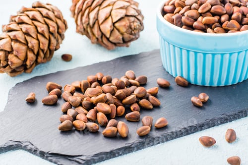Preview: A handful of unpeeled pine nuts on a slate board, pine cones and nuts in a bowl.