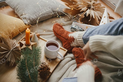 Preview: Woman legs in socks on blanket with cup of tea, christmas stars, lights, candle. Cozy home. Hygge