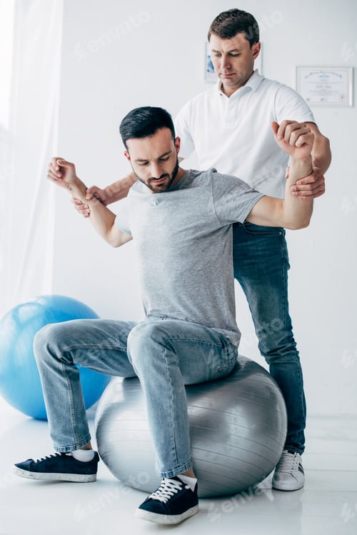 Preview: Physiotherapist stretching arm of handsome patient on fitness ball in hospital