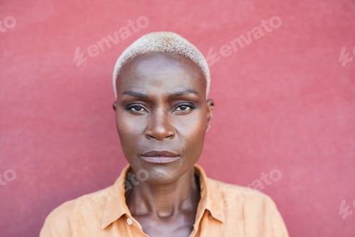 Preview: Portrait of mature african blond woman looking serious on camera with red background outdoor