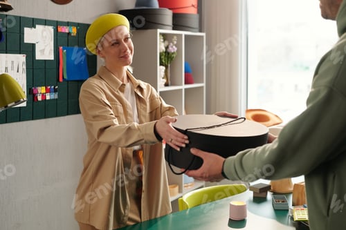 Preview: Happy young female shop assistant passing packed round box to buyer