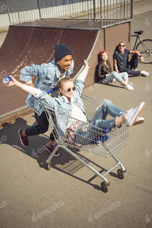 Preview: Boy carrying happy girl in shopping cart while friends sitting near ramp at skateboard park