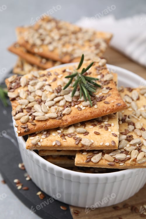 Preview: Cereal crackers with flax, sunflower, sesame seeds and rosemary in bowl on table, closeup