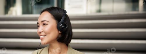 Preview: Smiling asian girl, laughing, listening music in headphones, sitting outdoors. Uni student enjoying