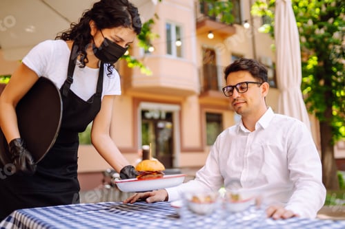 Preview: Waitress in protective mask serving delicious food for man in cozy restaurant.
