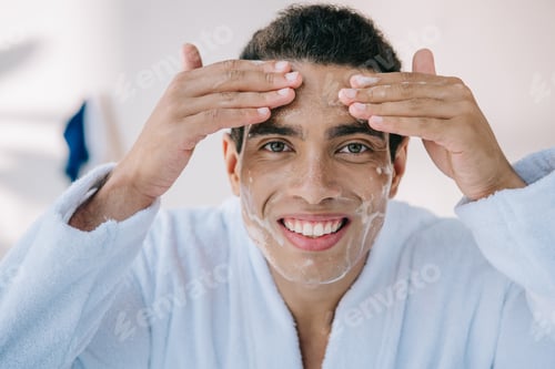 Preview: handsome man in bathrobe washing face with facial foam and smiling while looking at camera