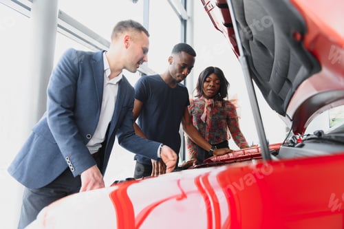 Preview: happy african car salesman showing car engine to customers
