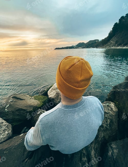 Preview: Caucasian guy in a blue sweater and hat holds his backpack and looking at the calm sea