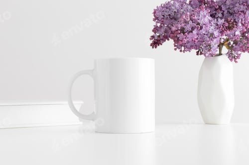 Preview: White mug mockup with a book and a bouquet of lilac in a vase on a white table.
