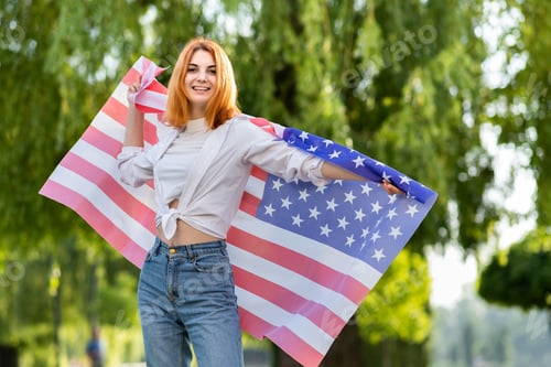 Preview: Young red haired woman holding USA national flag standing outdoors in summer park.