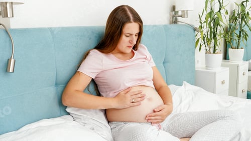 Preview: Young pregnant woman feeling unwell and abdominal pain sitting on bed. Healthcare and intoxication