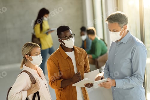 Preview: University professor and his students with face masks using digital tablet in a hallway.