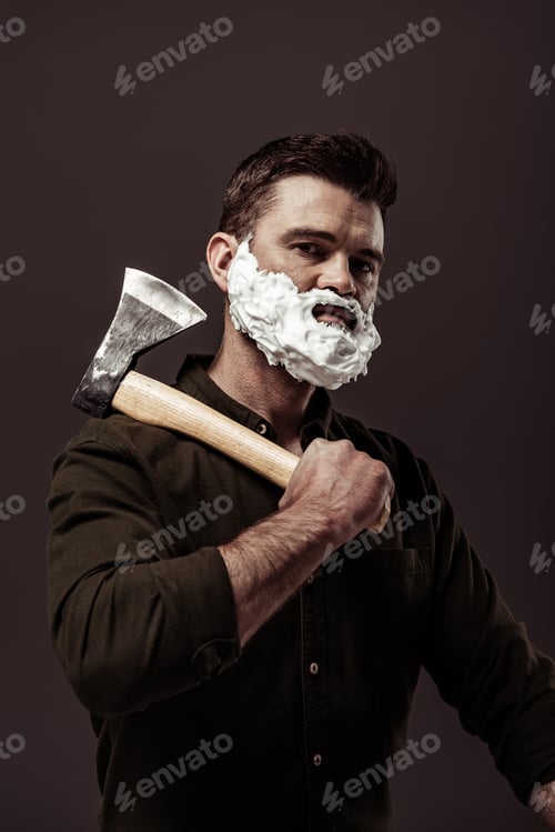Preview: handsome bearded man in brown shirt holding ax while looking at camera isolated on brown