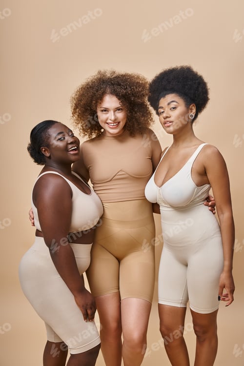 Preview: happy multiethnic plus size girlfriends in lingerie posing on beige backdrop, body positive beauty