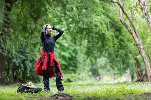 Preview: A young woman stands in a forest clearing with hands on her head, appearing thoughtful or tired.