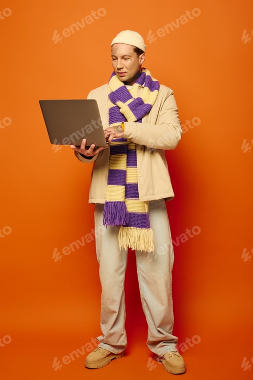 Preview: A young man with tattoos works on a laptop while standing against an orange backdrop
