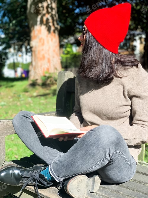 Preview: Young candid woman in casual autumnal clothes siting on a wooden bench in park and reading red book