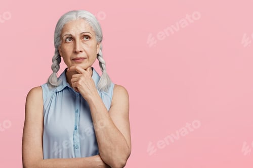 Preview: Pensive grey haired woman dressed in blouse, keeps hands on chin, looks upwards, isolated over pink