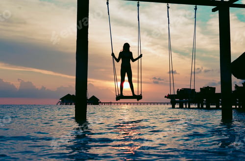 Preview: silhouette teen girl on a swing in the sea at sunset in the Maldives, the concept of travel