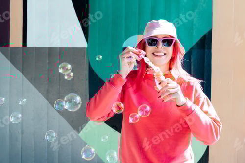 Preview: Playful smiling woman with pink hair, sunglasses in bucket hat blowing party soap bubbles